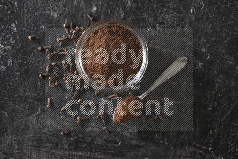 A glass bowl and a metal spoon full of cloves powder with gloves grains beside them on a textured black flooring