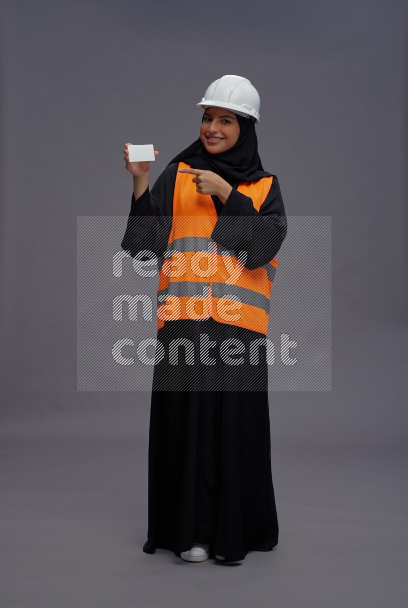 Saudi woman wearing Abaya with engineer vest standing holding ATM card on gray background