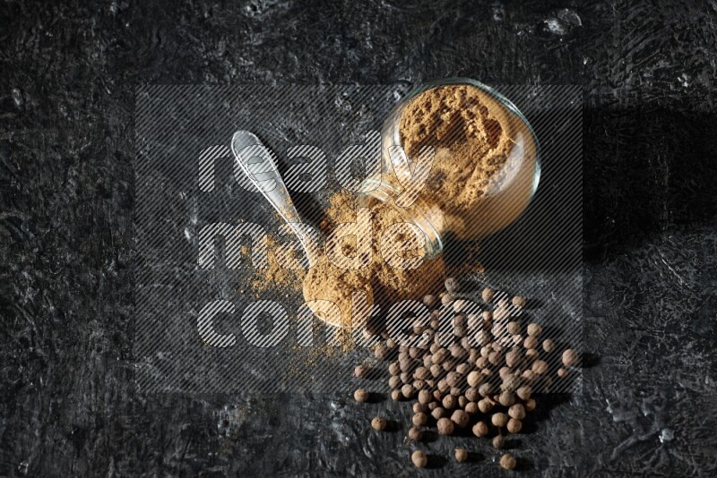 A flipped glass spice jar and metal spoon full of allspice powder and powder spilled out of it with whole balls on a textured black flooring
