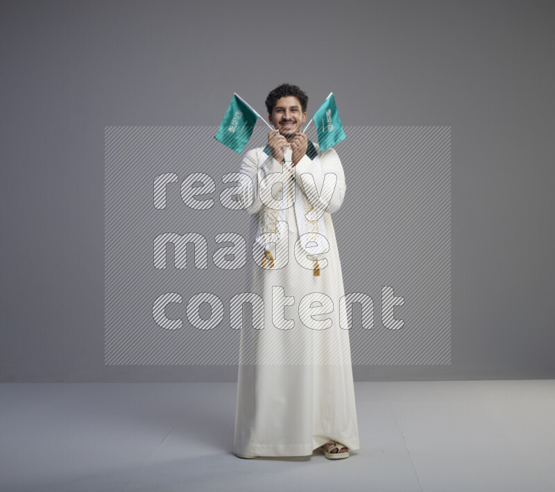 A Saudi man standing wearing thob and saudi flag scarf and holding small saudi flag on gray background