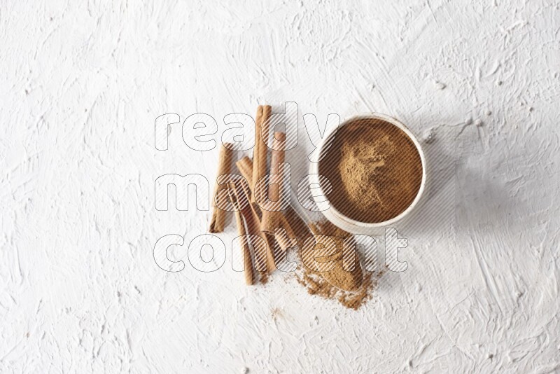 Ceramic beige bowl full of cinnamon powder and a metal spoon with cinnamon sticks next of it on a textured white background