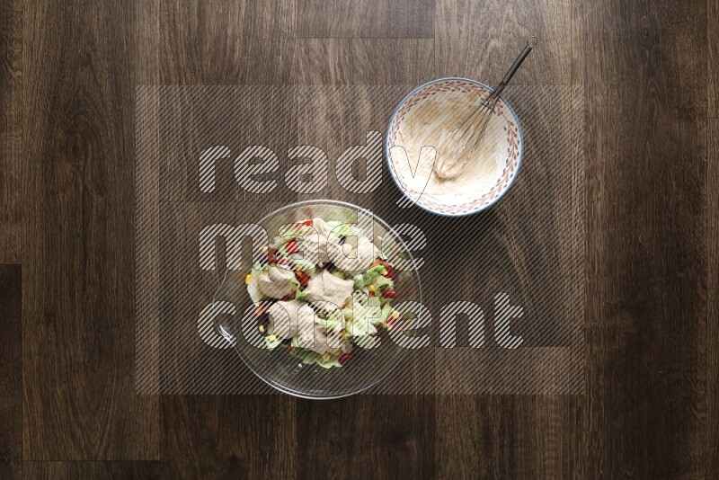 A bowl full of salad (avocado, tomatoes, red beans, olives, bell pepper, corn, lettuce) and bowl of salad dressing on wooden background