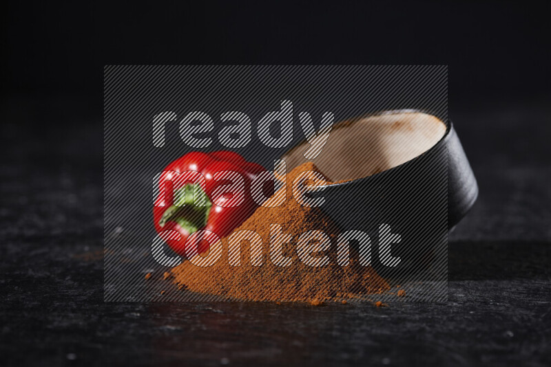 A black pottery bowl full of ground paprika powder with fallen powder from it on black background