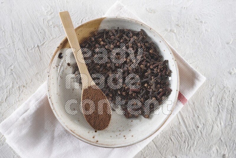 A Pottery plate full of whole cloves and a wooden spoon full of cloves powder in it on a textured white background