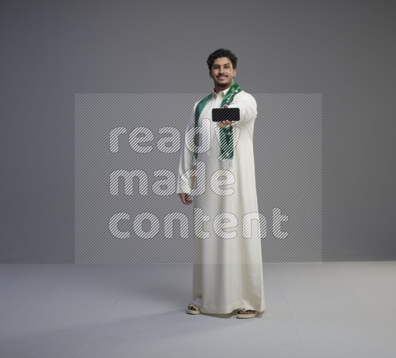 A Saudi man standing wearing thob and Saudi flag scarf showing phone to camera on gray background