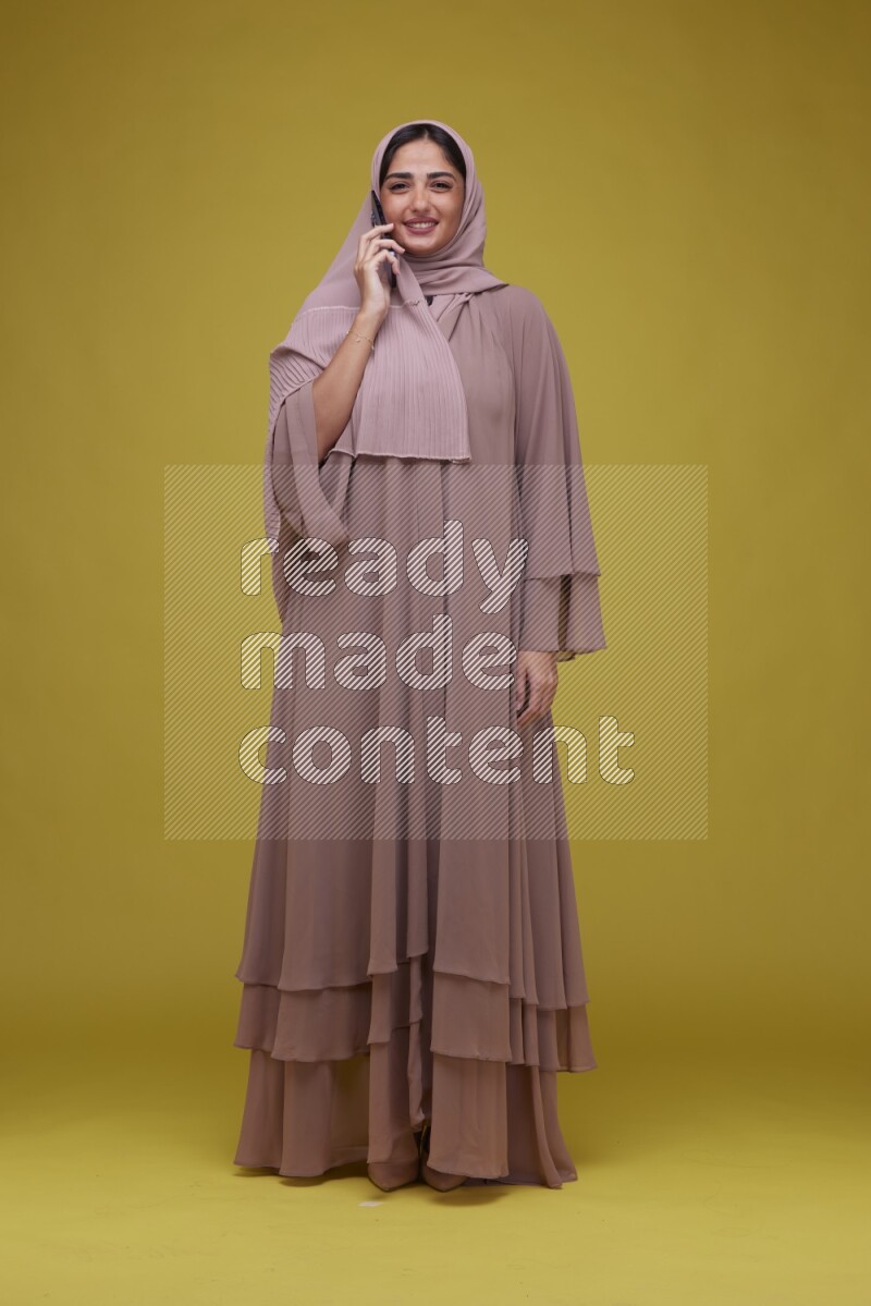 A woman having a call on a Yellow Background wearing Brown Abaya with Hijab