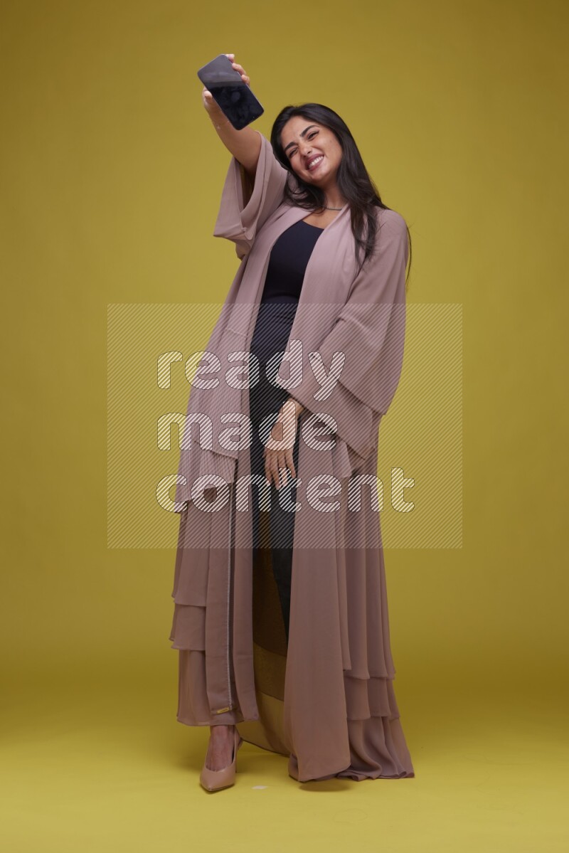A woman Showing SmartPhone Screen on a Yellow Background wearing Brown Abaya