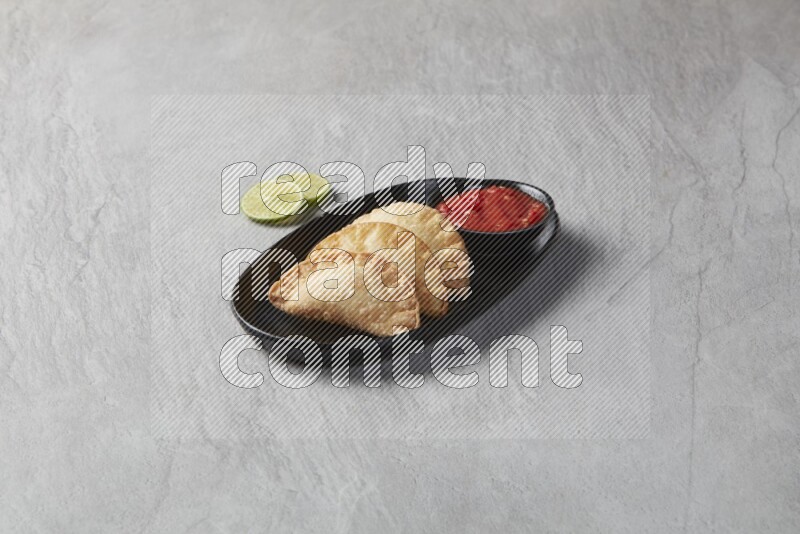 Three fried sambosas in an oval shaped black plate and a red sauce in a black round ramekin on a gray background