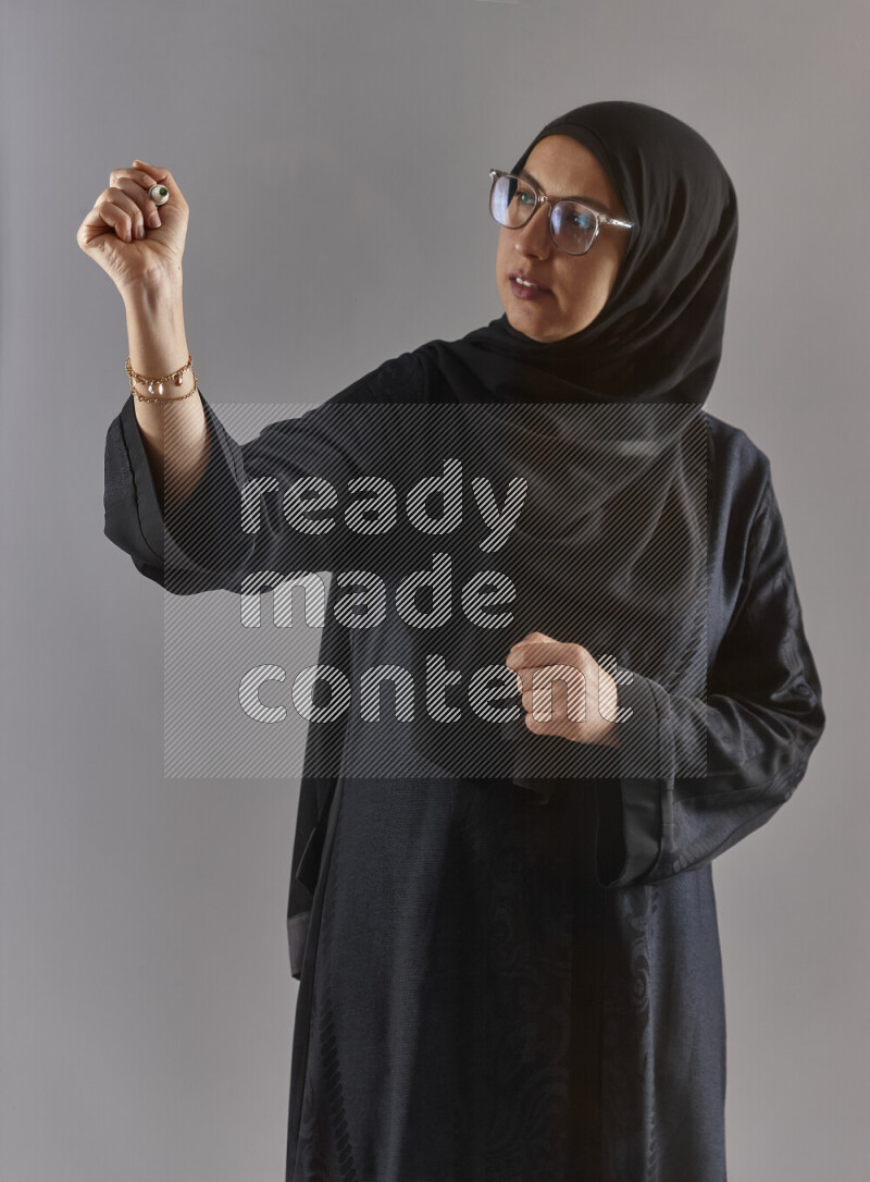 Woman in black abaya, hijab and glasses holding a marker pen to write on a transparent board with different reactions and expressions on grey background