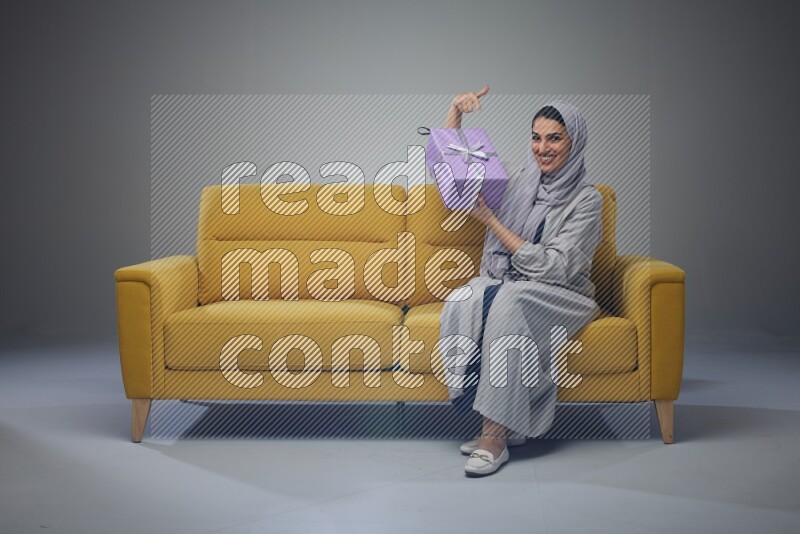 A Saudi woman wearing a light gray Abaya and head scarf sitting on a yellow sofa holding a gift box beside multi color gift boxes eye level on a grey background