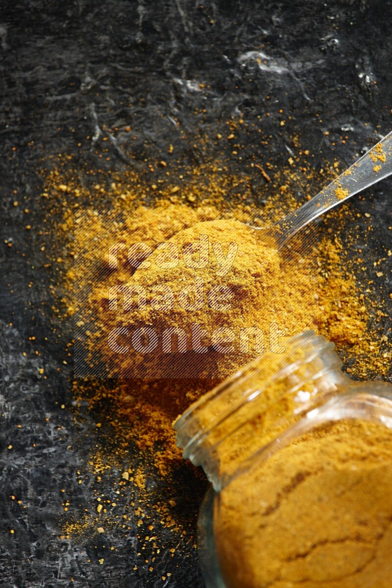 A flipped glass spice jar and a metal spoon full of turmeric powder and powder spilled out of it on textured black flooring
