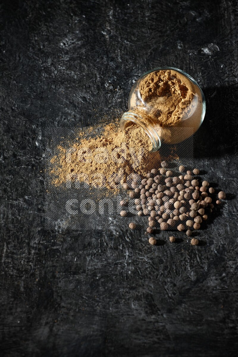 A flipped glass spice jar full of allspice powder and powder spilled out of it with whole balls on a textured black flooring