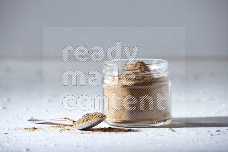 A glass jar and a metal spoon full of allspice powder on a textured white flooring