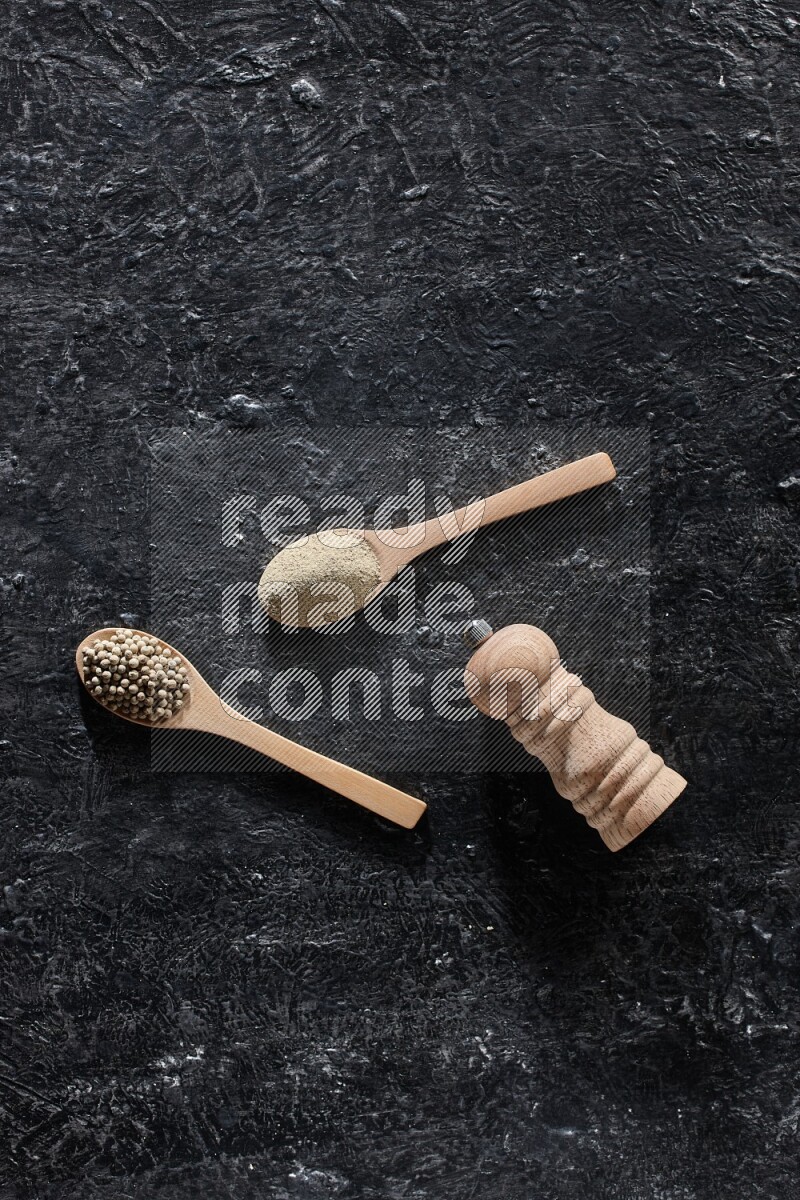 2 wooden spoons one full of white pepper powder and the other with pepper beads and a wooden pepper mill on textured black flooring