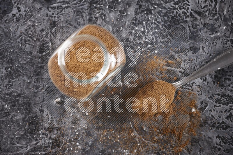 Herbal glass jar full of cinnamon powder and a metal spoon full of powder on textured black background