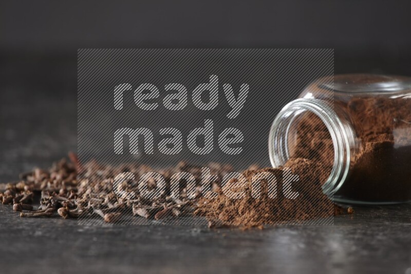 A flipped glass spice jar full of cloves powder and powder came out of it with cloves spread on textured black flooring