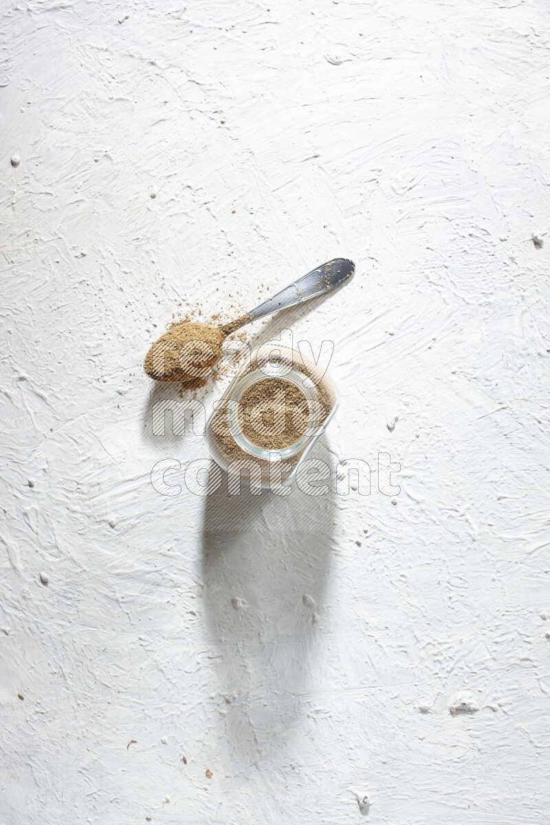 وعاء زجاجي للتوابل وملعقة معدنية ممتلئان ببودرة الكمون على خلفية بيضاء