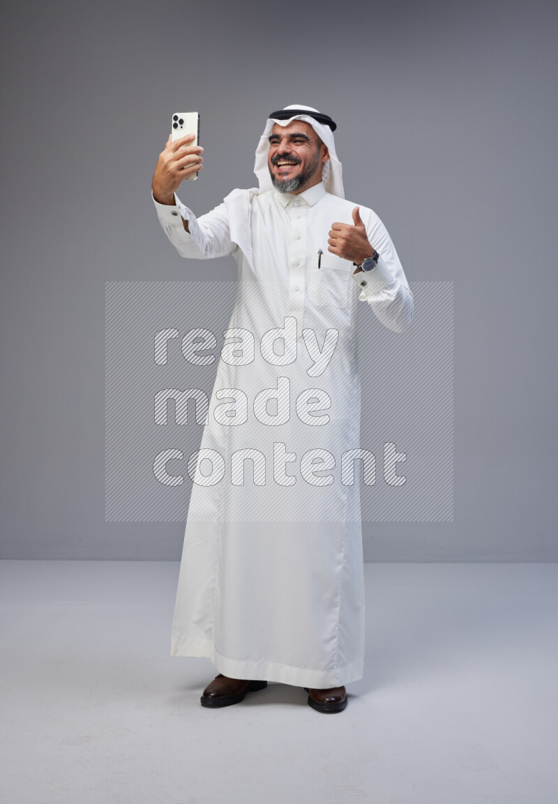 Saudi man Wearing Thob and white Shomag standing taking selfie on Gray background