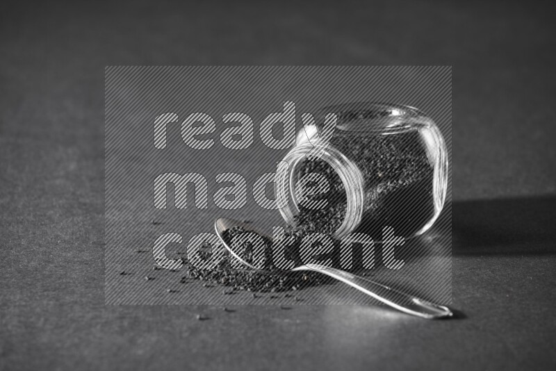A flipped glass spice jar full of black seeds and the seeds spread out with a metal spoon full it on a black flooring