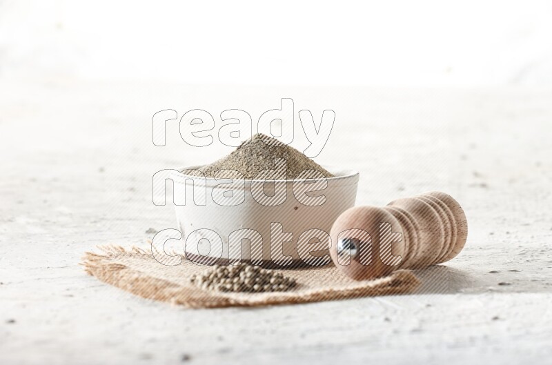 White pottery bowl full of white pepper powder set on a burlap piece of fabric with pepper beads and wooden pepper grinder on textured white flooring