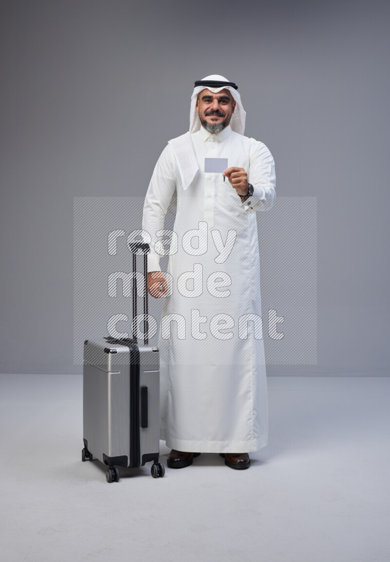 Saudi man wearing Thob and white Shomag standing holding Travel bag and ATM card on Gray background