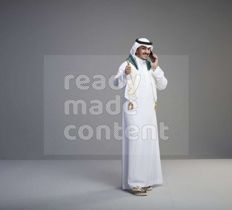 A saudi man standing wearing thob and white shomag with flag scarf on his neck talking on phone on gray background