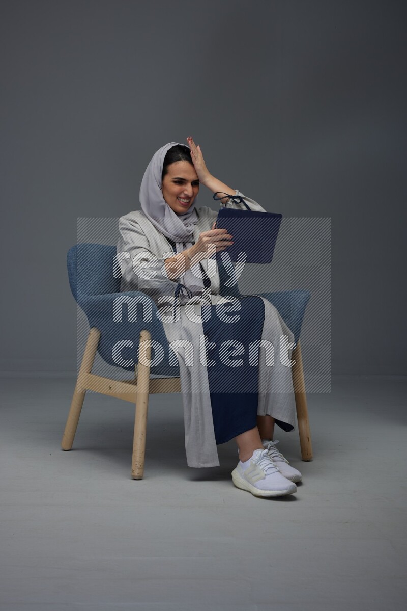 A Saudi female wearing a light gray Abaya and head scarf sitting on a dark grey chair while holding her iPad on a grey background