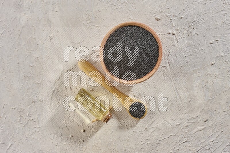A wooden bowl and spoon full of black seeds with a bottle of black seeds oil on a textured white flooring