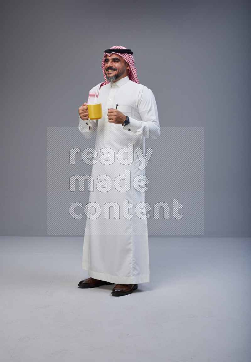 Saudi man Wearing Thob and red Shomag standing holding mug on Gray background