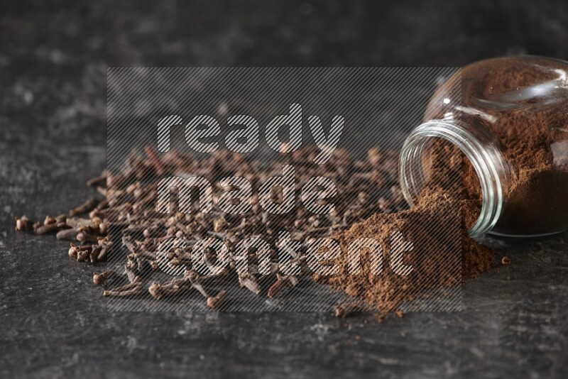 A flipped glass spice jar full of cloves powder and powder came out of it with cloves spread on textured black flooring