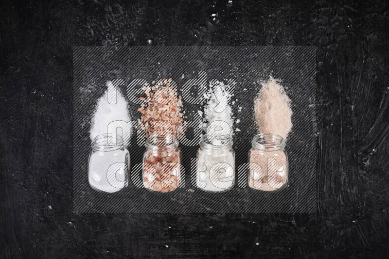 4 glass jars filled with table salt, coarse sea salt, fine himalayan salt and coarse himalayan salt on black background