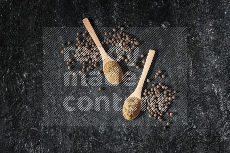 Wooden spoons full of all spice powder and allspice whole balls beside it on a textured black flooring