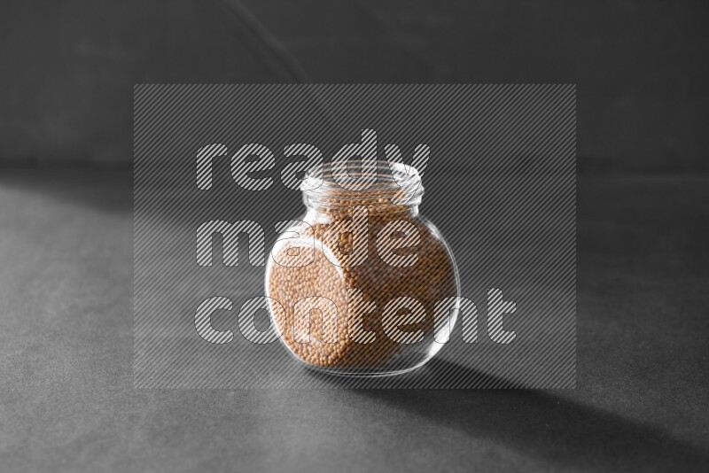 A glass spice jar full of mustard seeds on black flooring