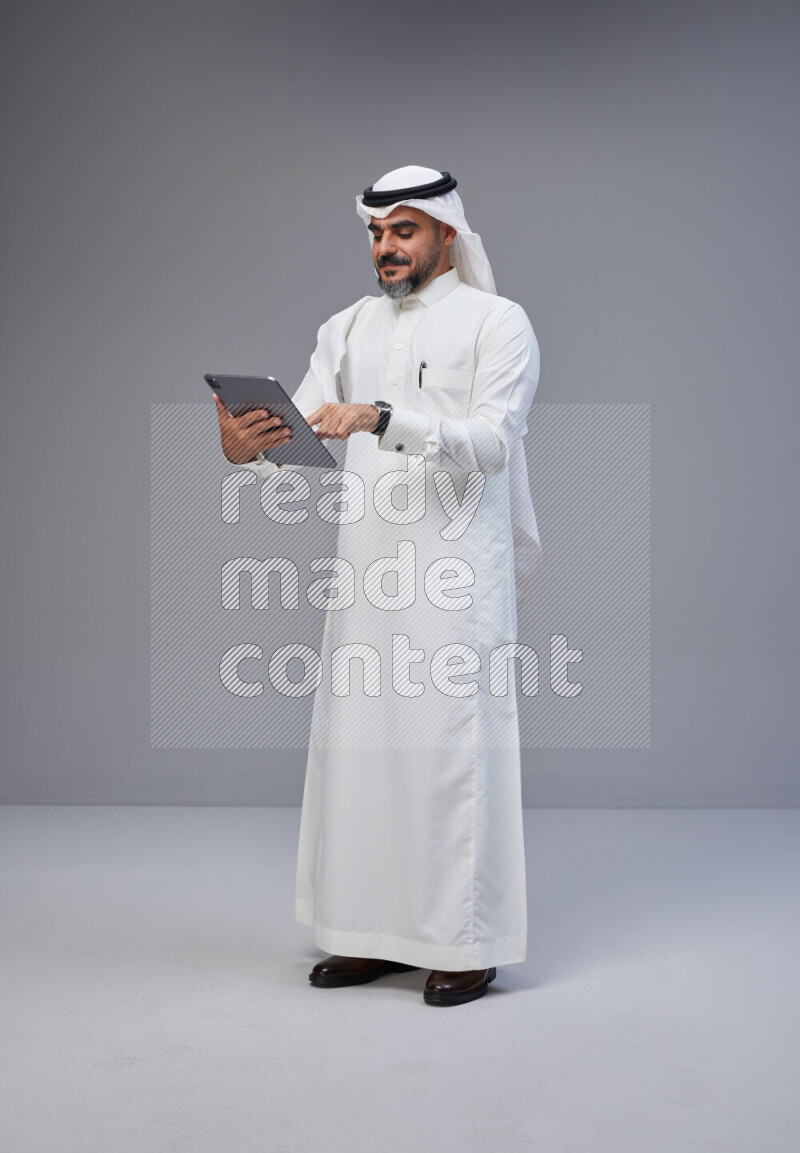 Saudi man Wearing Thob and white Shomag standing working on tablet on Gray background
