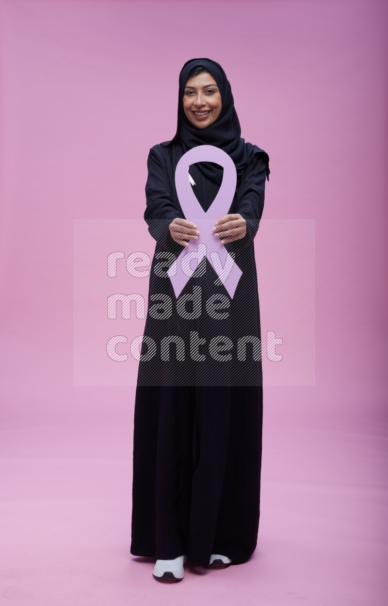 Saudi woman wearing pink ribbon on abaya standing holding awareness ribbon on plain pink background