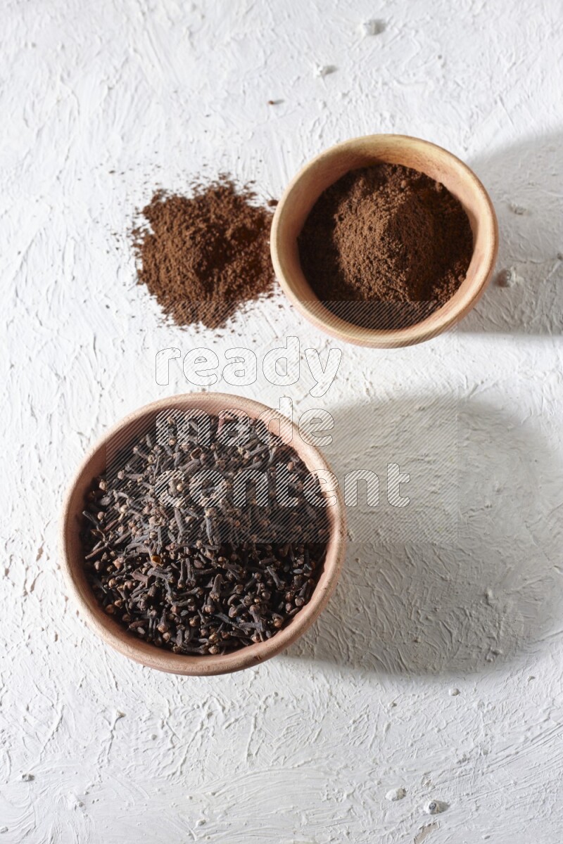 2 wooden bowls full of cloves powder and whole cloves on a textured white flooring