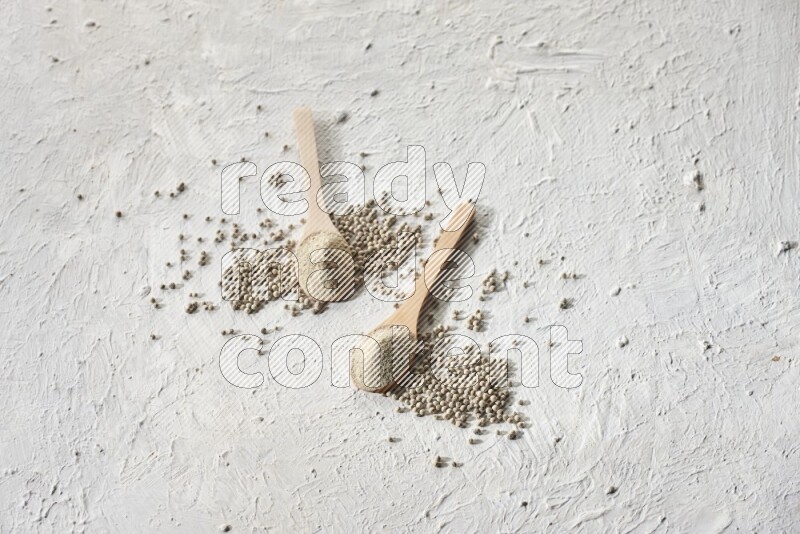 2 wooden spoons full of white pepper powder with white pepper beads on textured white flooring