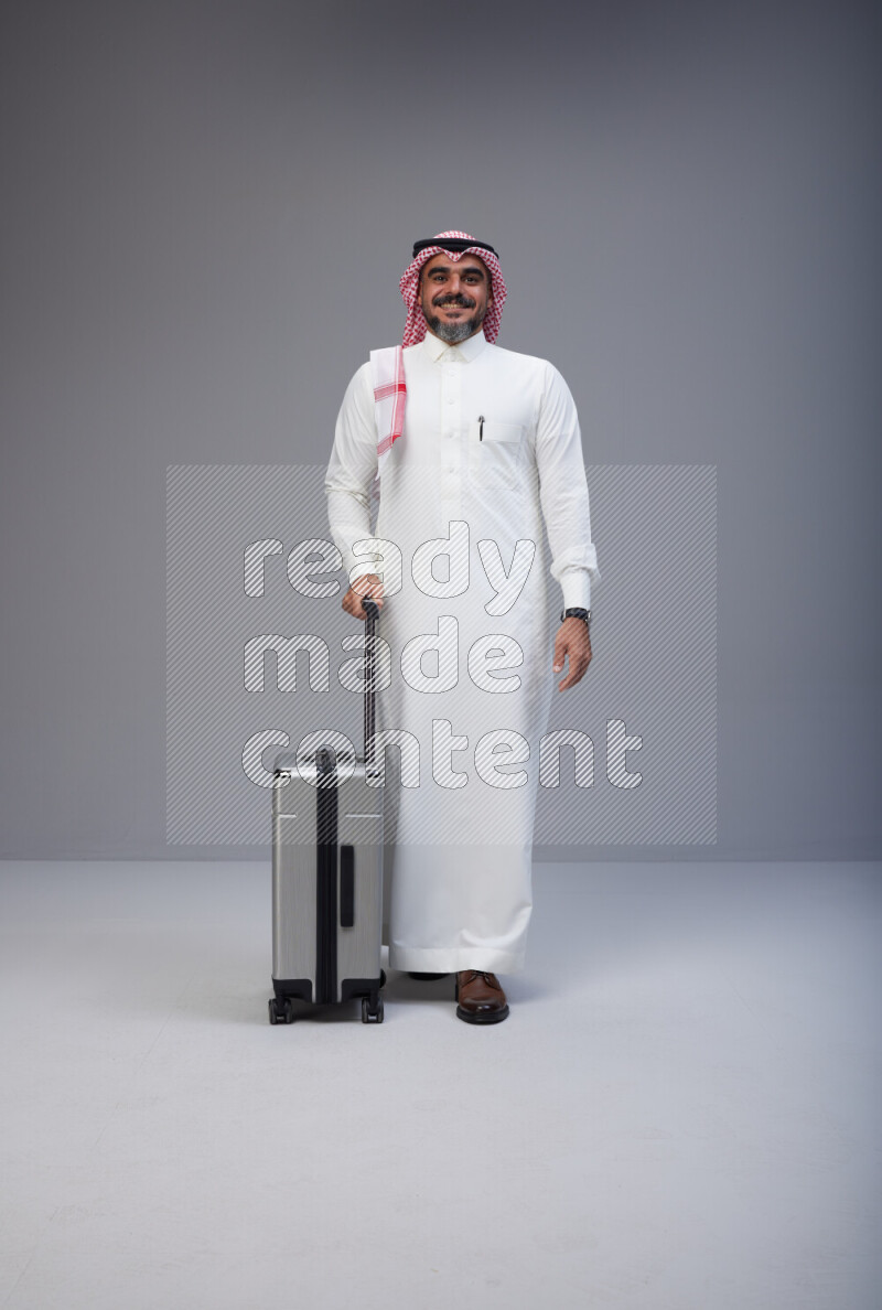 Saudi man wearing Thob and red Shomag standing holding Travel bag on Gray background