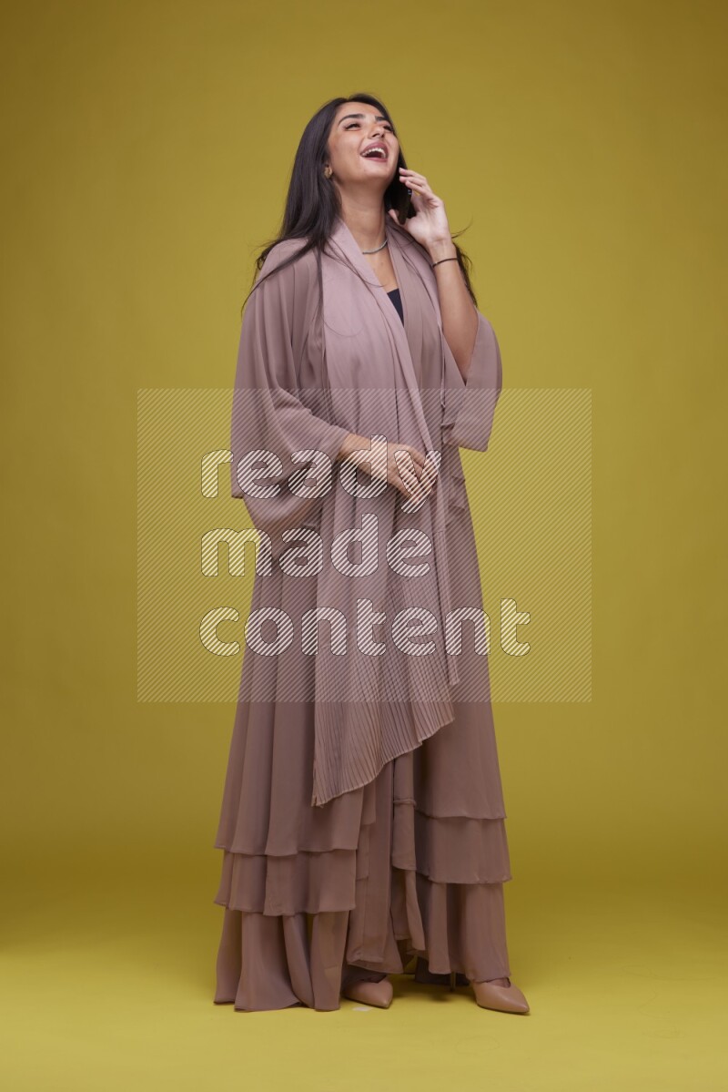 A woman Calling on a Yellow Background wearing Brown Abaya
