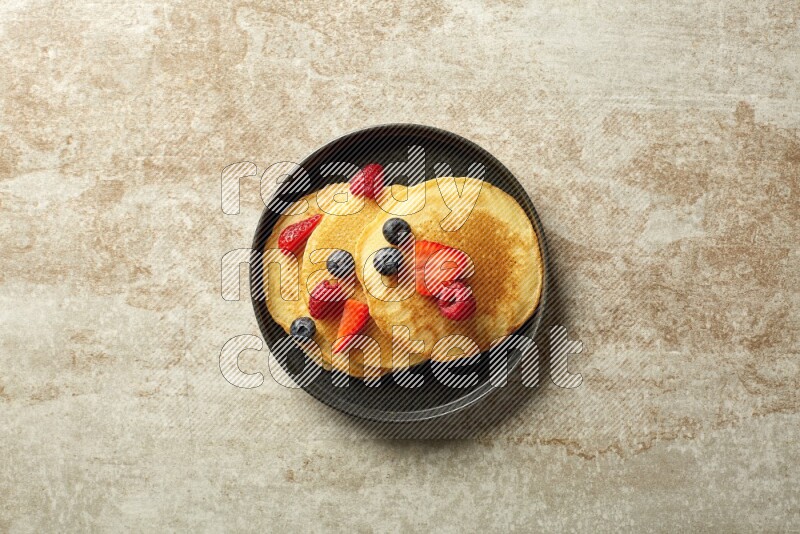 Three stacked mixed berries pancakes in a black plate on beige background