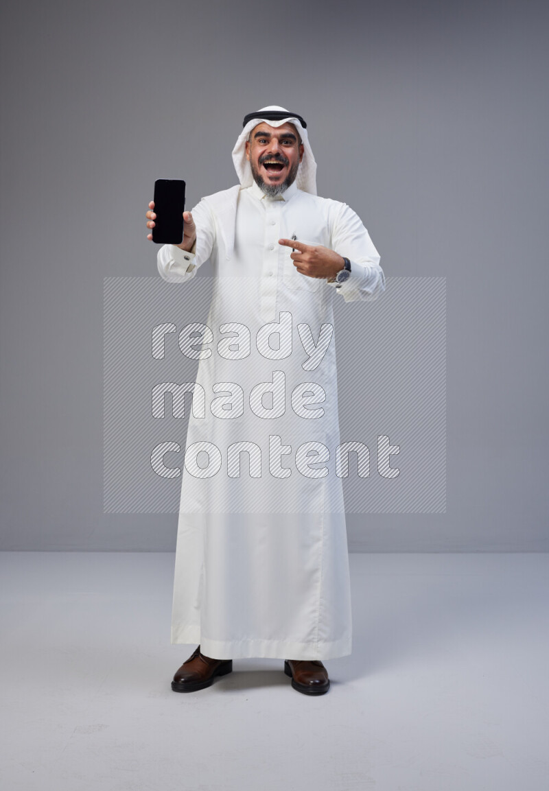 Saudi man Wearing Thob and white Shomag standing showing phone to camera on Gray background