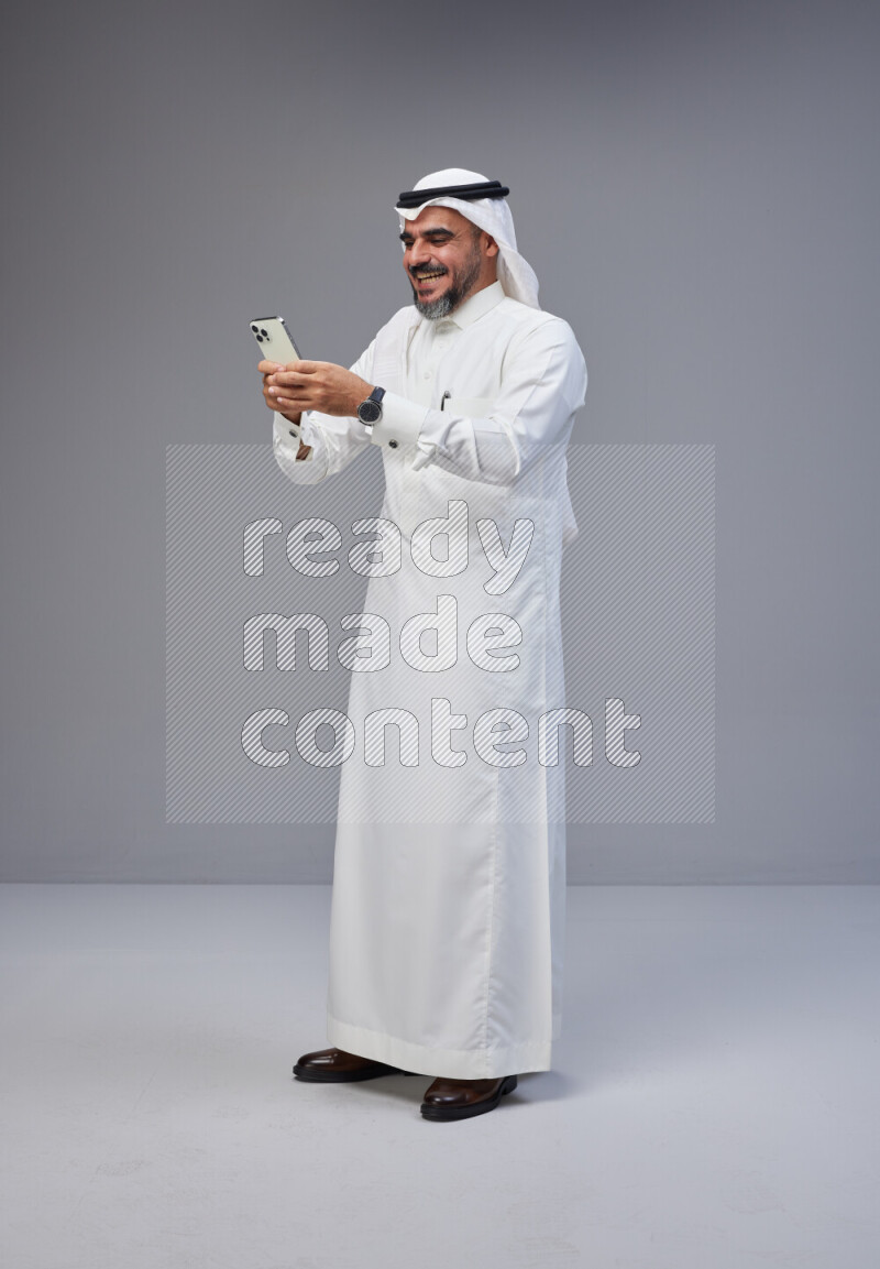 Saudi man Wearing Thob and white Shomag standing texting on phone on Gray background