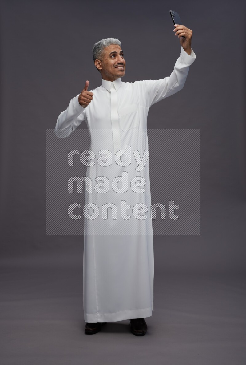 Saudi man wearing thob standing taking selfie on gray background