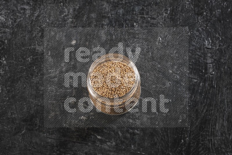 A glass jar full of mustard seeds on a textured black flooring
