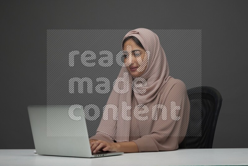 A Saudi woman Setting on her desk Typing on her laptop on a Gray Background wearing Brown Abaya with Hijab