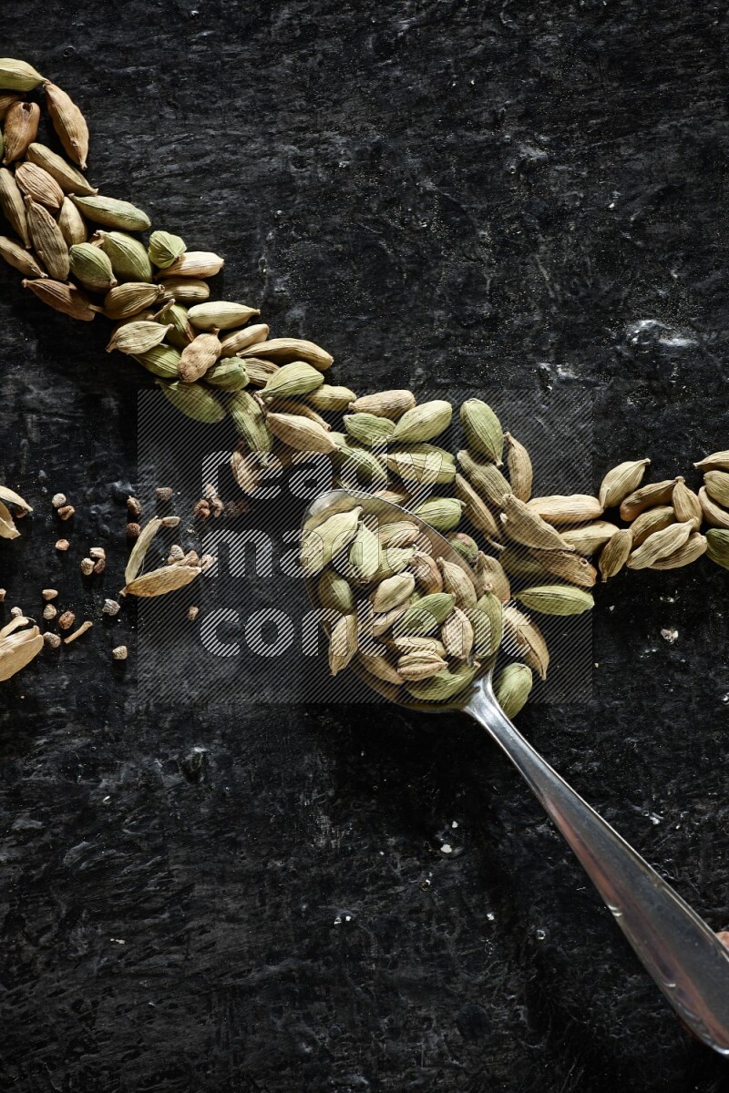 A Metal spoon full of cardamom seeds and some seeds beside it on a textured black flooring