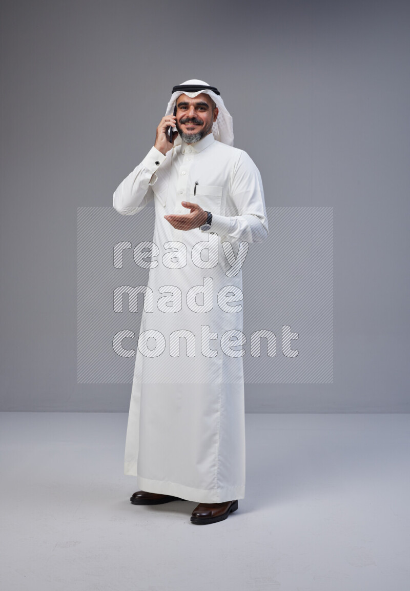 Saudi man Wearing Thob and white Shomag standing talking on phone on Gray background