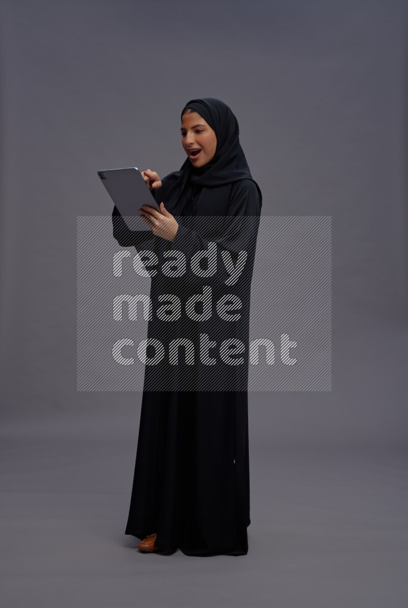 Saudi woman wearing Abaya standing working on tablet on gray background