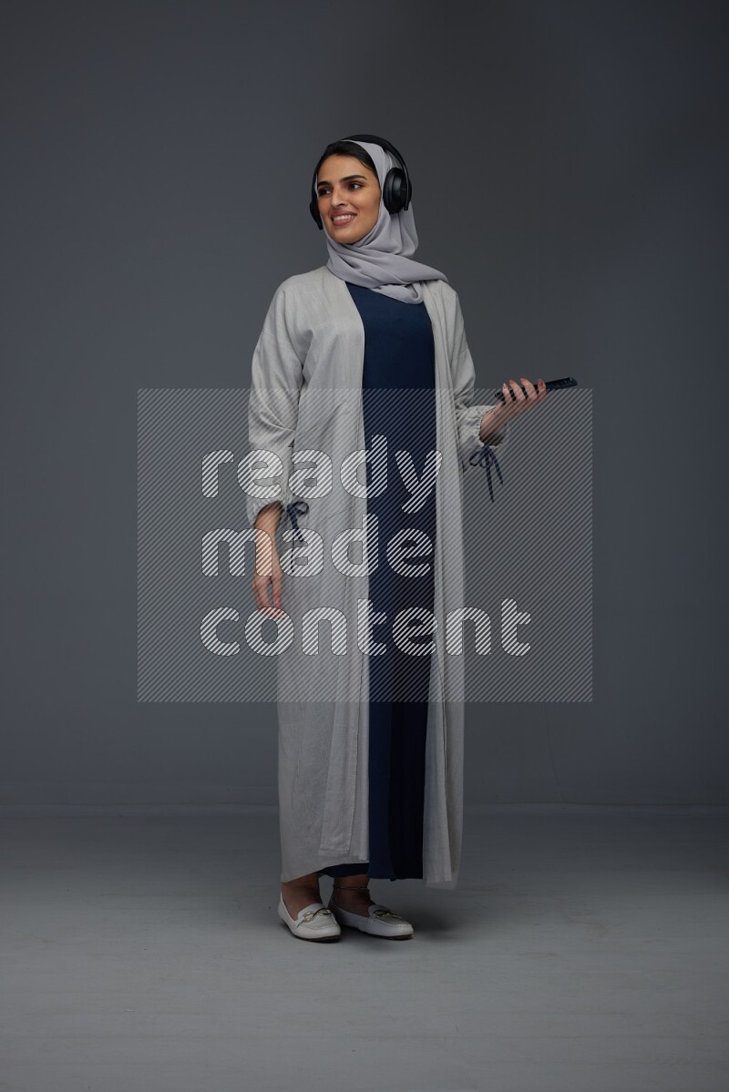 A Saudi woman wearing a light gray Abaya and head scarf standing and listening to music on a grey background