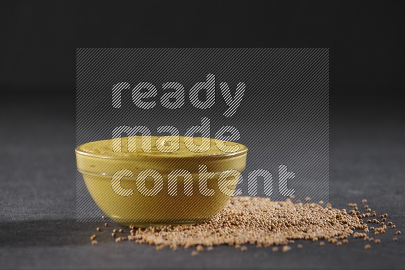A glass bowl full of mustard paste with mustard seeds underneath on black flooring